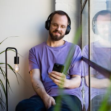 Ein Mann mit Kopfhörern, sitzt in der Fensterbank und schaut auf sein Smartphone.