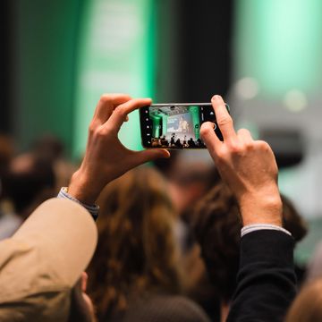 Eine Mann hält sein Smartphone in die Höhe und fotografiert eine Veranstaltung.